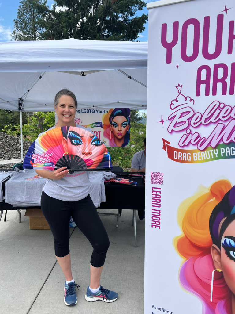 Believe in Me volunteer smiling at Spokane Pride 2023 booth while holding a rainbow drag-queen clack fan in support of LGBTQIA2S+ youth
