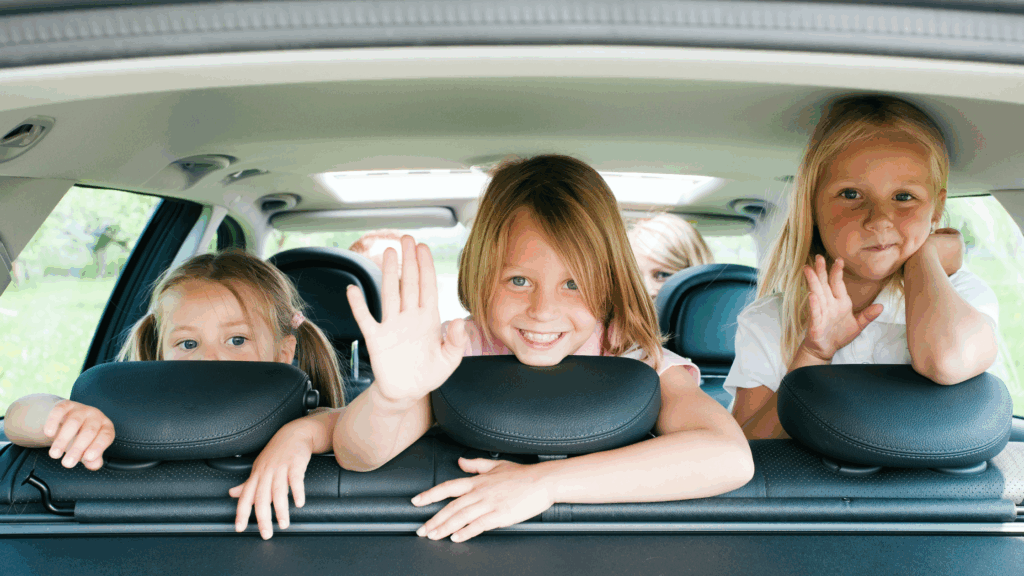 Three girls peek over car headrests; the center child smiles and waves from the back seat.