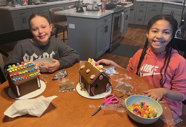 Two participants decorate gingerbread houses with colorful candy at a table.